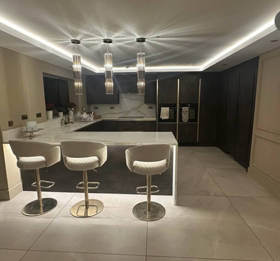 Modern fitted kitchen with a large island, three white bar stools, pendant lights, and dark cabinetry.