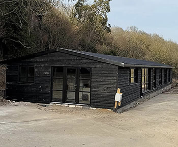 A long, dark-colored wooden building with multiple windows, situated on a dirt surface with trees in the background.