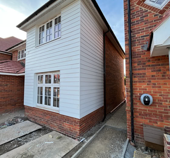 A narrow alley between two houses, one with white siding and the other with red brick. The ground is partially paved, and there are windows on both houses.