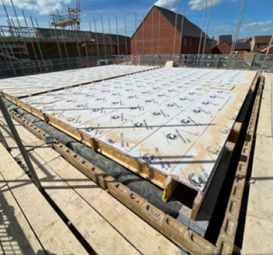 A construction site with a large concrete slab in progress, surrounded by scaffolding and wooden beams, with brick buildings and a blue sky with clouds in the background.