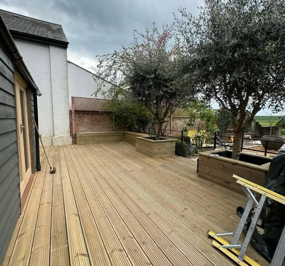 Wooden deck with trees and plants along the side, adjacent to a building with dark siding and a white wall.