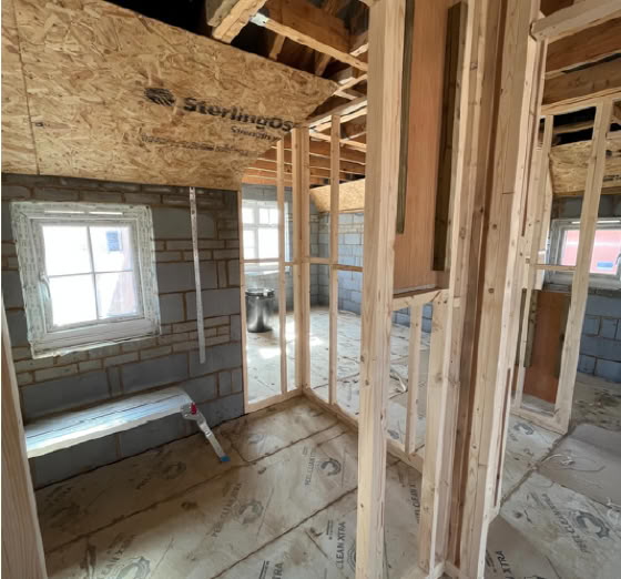 Interior view of a building under construction, showing wooden framing, exposed beams, and a partially completed wall with a window.