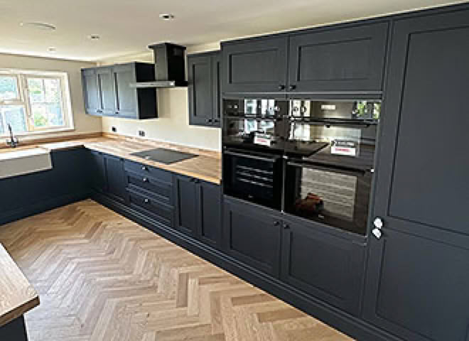 Modern kitchen fitter installation with dark cabinets, built-in ovens, a light wood herringbone floor, and large windows.