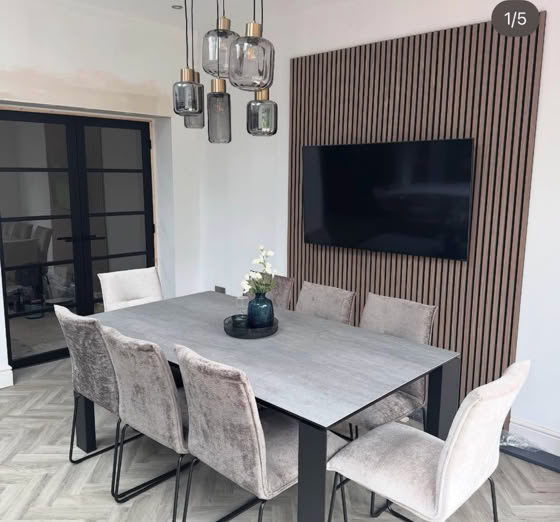 A modern dining room with a rectangular table surrounded by six upholstered chairs, a wall-mounted TV, pendant lights above, and a decorative wall panel.