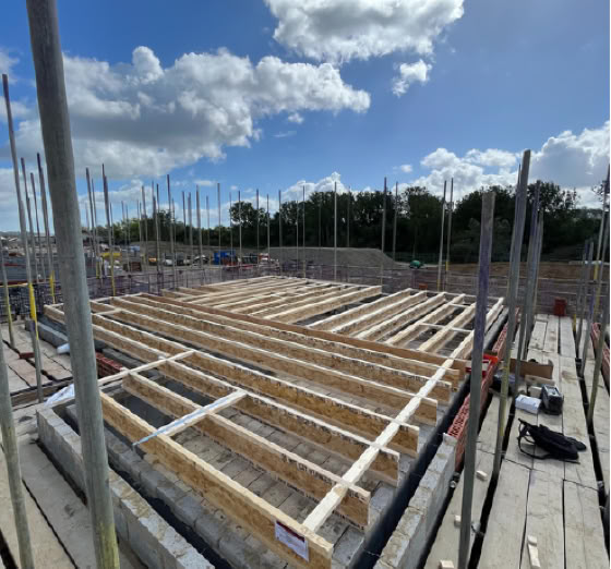 Construction site with wooden framework on a concrete foundation, surrounded by scaffolding poles, under a partly cloudy sky.
