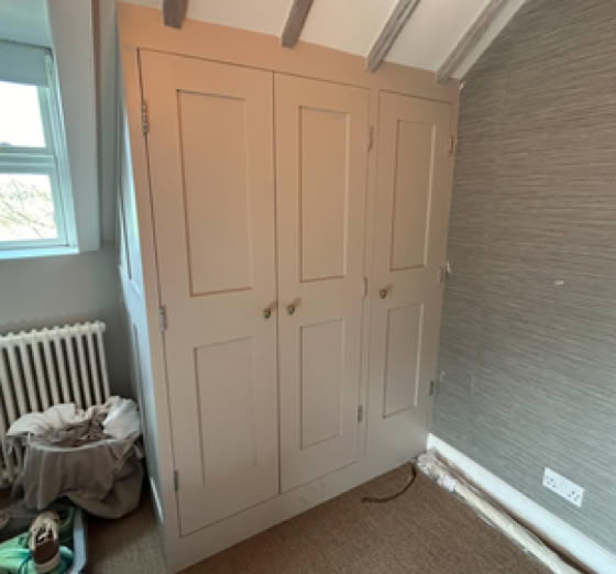 A room with a beige built-in wardrobe, a window with a radiator beneath it, and a basket with clothes on the floor.