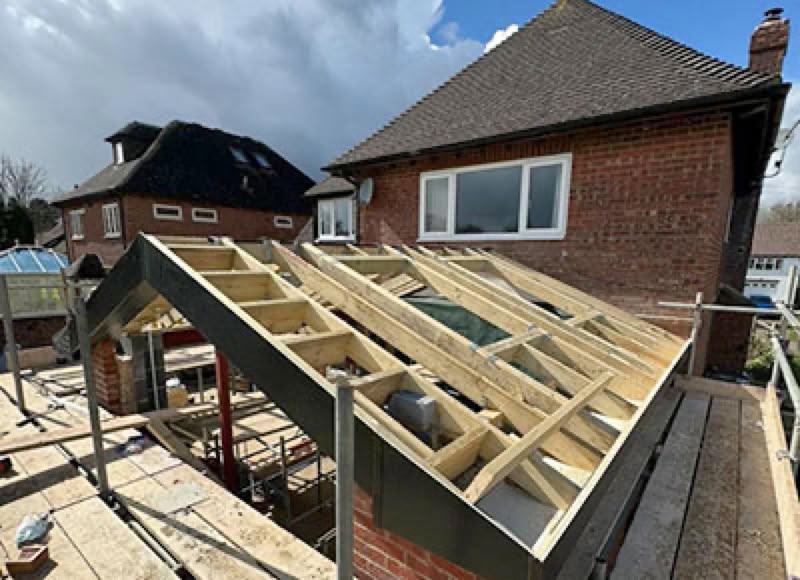A partially constructed wooden roof frame on a brick building, with scaffolding around it and another house visible in the background under a cloudy sky.
