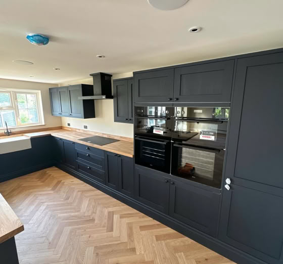 Modern kitchen with dark cabinets, built-in ovens, a light wood herringbone floor, and large windows.