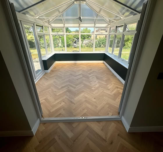 View of an empty sunroom with glass walls and ceiling, featuring a herringbone wood floor pattern, and a garden visible outside.